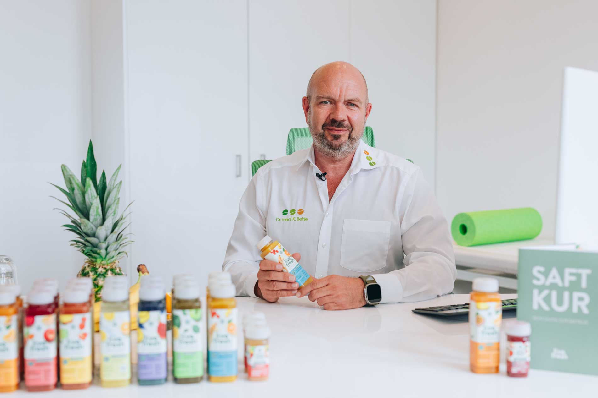 Mann im weißen Hemd mit Aufschrift „Dr. med K. Behrle“ sitzt an einem Schreibtisch und hält eine Flasche Saft. Vor ihm stehen verschiedene Säfte, im Hintergrund eine Ananas, eine grüne Yogamatte und eine grüne Broschüre mit der Aufschrift „Saftkur“.