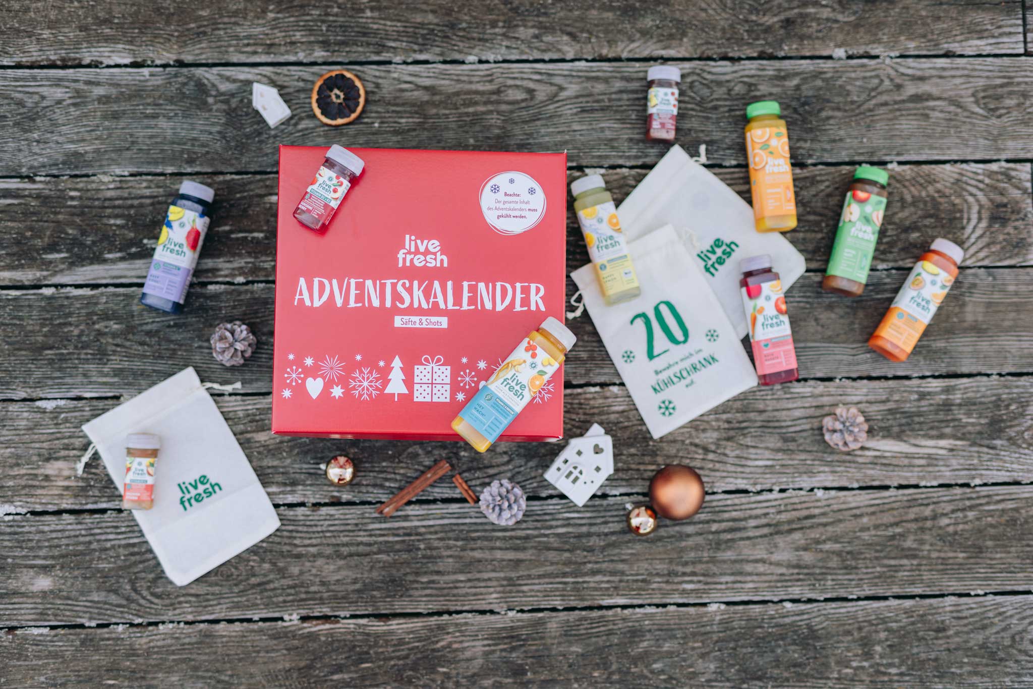 LiveFresh Adventskalender in einer roten Box mit winterlichen Motiven, umgeben von verschiedenen Saft- und Shot-Flaschen auf einer rustikalen Holzoberfläche. Mehrere weiße Stoffbeutel mit LiveFresh-Logo und einer Zahl liegen daneben. Dekoriert mit Tannenzapfen, Zimtstangen, getrockneten Orangenscheiben und Weihnachtskugeln.