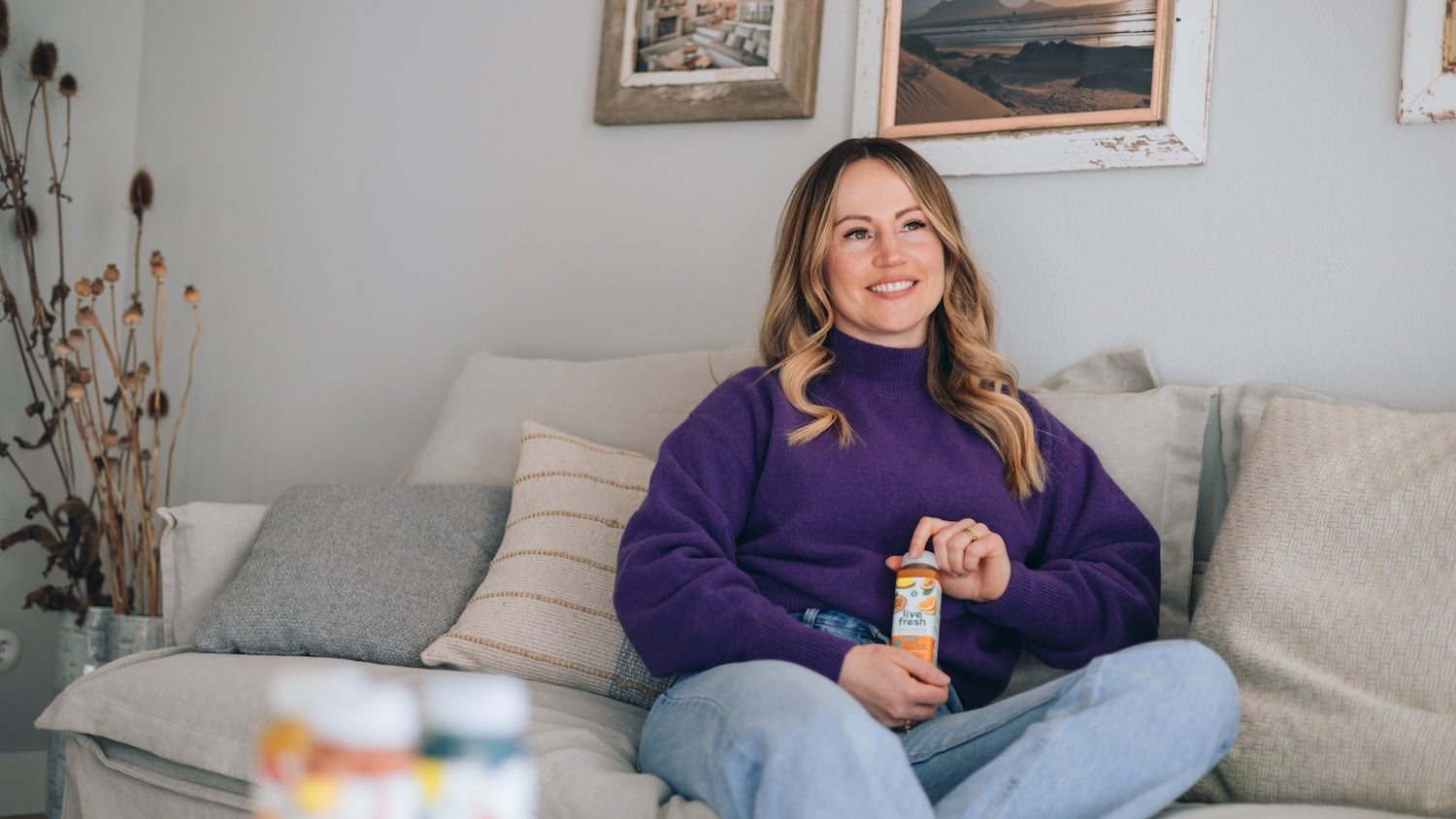 Lächelnde Frau in einem lila Pullover und hellblauer Jeans entspannt auf einem beigen Sofa, hält eine LiveFresh-Shotflasche in der Hand. Gerahmte Fotos und Trockenblumen schmücken den gemütlichen Wohnraum.