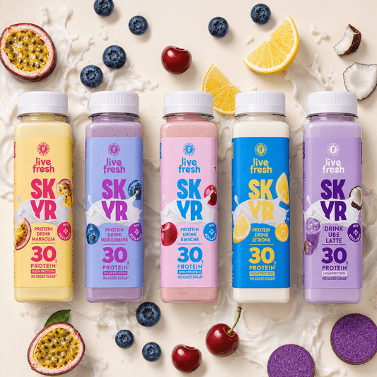 Fünf bunte Flaschen Skyr Proteindrink von Live Fresh in den Sorten Maracuja. Heidelbeere. Kirsche. Zitrone und Ube Latte. Rund um die Skyr Drinks liegen passende Früchte wie Maracuja. Heidelbeeren. Kirschen. Zitronen und Kokosnuss in Milchspritzern.
