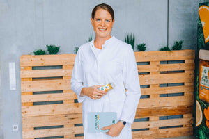 Eine lächelnde Frau mit braunen, zusammengebundenen Haaren in einem weißen Kittel hält eine Flasche Saft von LiveFresh in der einen Hand und ein Buch mit der Aufschrift "FOOD KUR" in der anderen Hand. Im Hintergrund stehen Pflanzen auf Holzpaletten vor einer grauen Wand.