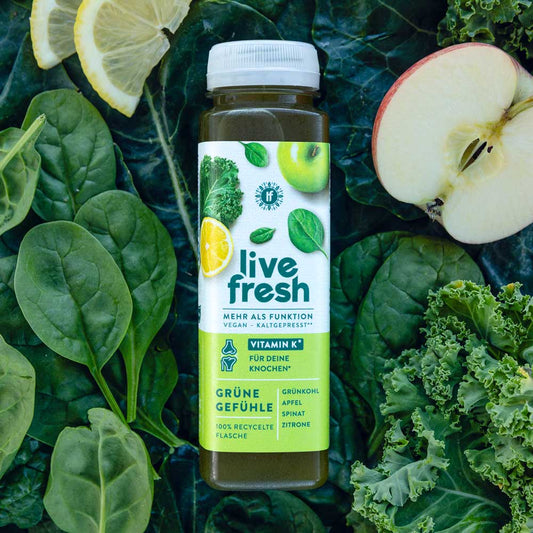 „Live Fresh“ Grüner Saft „Grüne Gefühle“ mit Vitamin K, umgeben von Spinat, Grünkohl, Apfel und Zitrone auf dunklem Blattgemüse-Hintergrund.