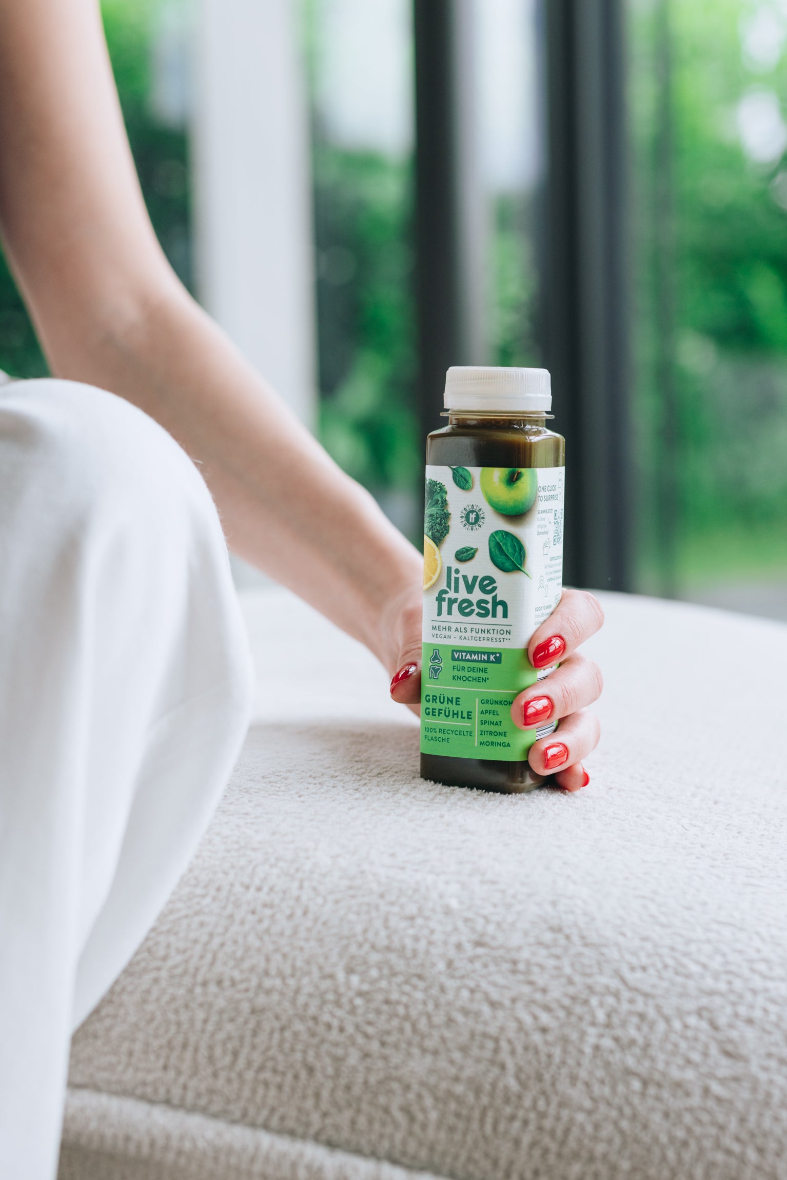 Eine Frau, die sitzend den Saft Grüne Gefühle von LiveFresh auf einem hellen Sofa hält. Es ist nur ihre Hand, mit roten Fingernägeln, und ihre Knie, mit einer weißen Hose bedeckt, zu sehen. Hintergrund ist verschwommen.