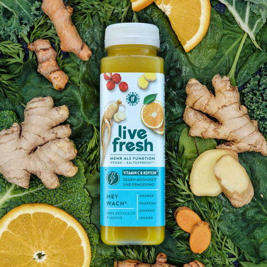 Eine Flasche „LiveFresh Hey Wach“ Saft, umgeben von frischen Zutaten wie Ingwer, Kurkuma, Orangen und Spinatblättern. Der Saft ist vegan, kaltgepresst und enthält Vitamin C sowie Koffein. Zu den Hauptzutaten gehören Orange, Guarana, Ginseng und Ingwer. Die Flasche besteht aus 100% recyceltem Material.