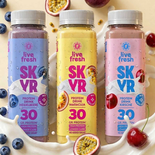 Drie flessen Skyr Proteïnedrank met bosbes, passievrucht en kers staan voor een romige melkplas, omringd door bijpassend fruit. Op de etiketten staat "live fresh", "SKYR", "PROTEIN DRINK BLUEBERRY", "PROTEIN DRINK MARACUJA", "PROTEIN DRINK CHERRY", "WITH COLLAGEN", "30 GR. PROTEIN", "HIGH PROTEIN", "LOWER CARB" en "NO ADDED SUGAR".
