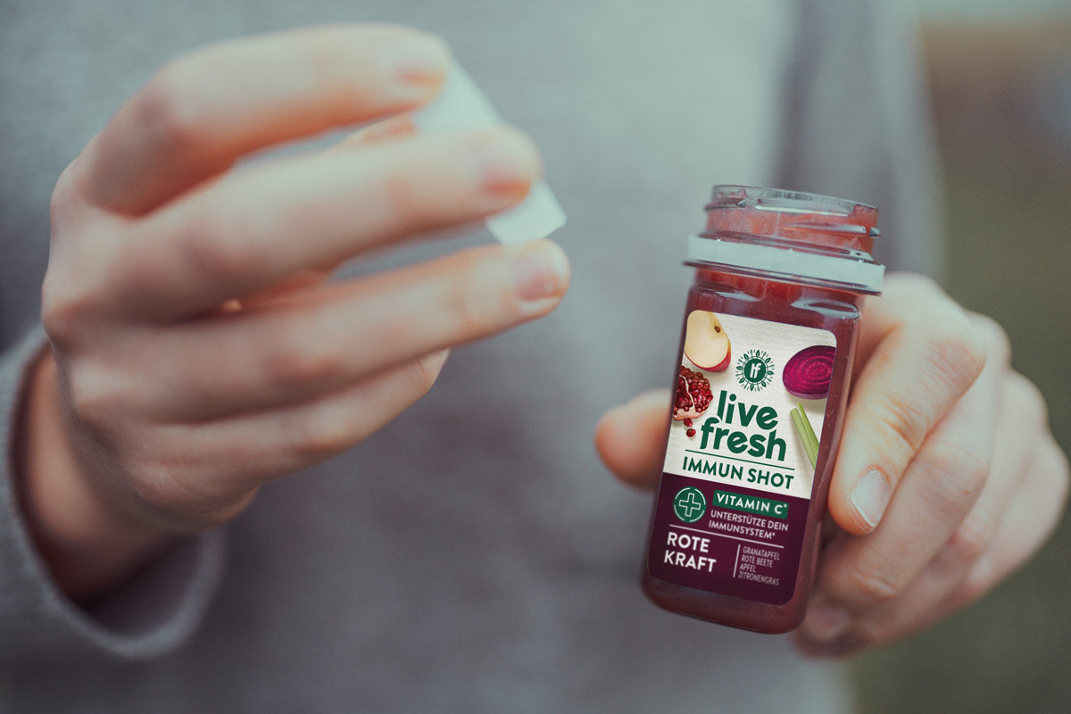 Eine Person in grauer Kleidung hält einen geöffneten Shot namens "Rote Kraft" von LiveFresh in der Hand und nimmt den Deckel ab. Das Etikett zeigt Zutaten wie Granatapfel, Rote Beete, Apfel und Zitronengras.