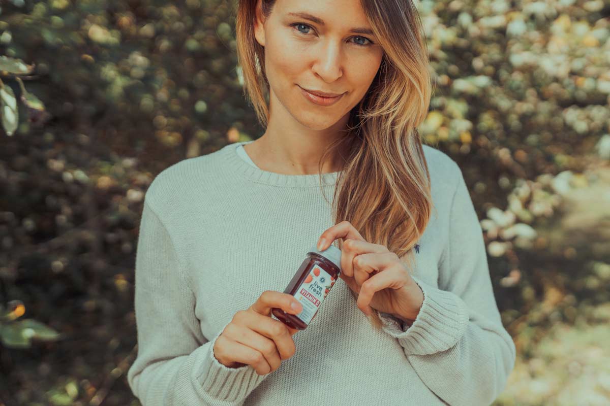 Eine lächelnde Frau mit langem, blonden Haar trägt einen hellgrauen Pullover und hält eine kleine Flasche "LiveFresh" Vitamin Shot in der Hand. Der Hintergrund zeigt eine natürliche, grüne Umgebung.