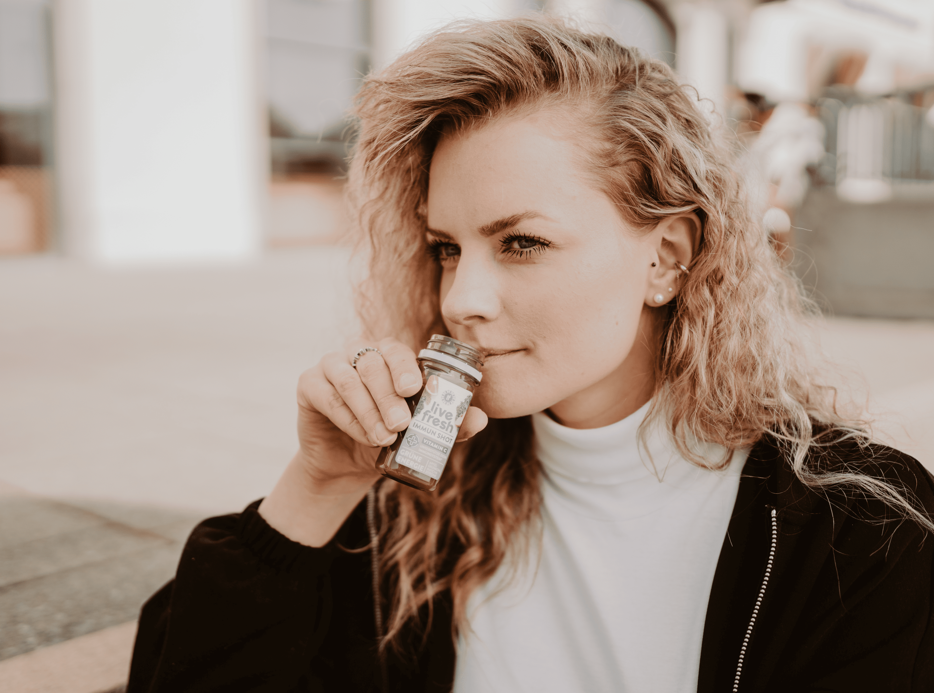 Frau mit lockigem, blondem Haar hält einen Live Fresh Immun Shot an ihre Lippen, bereit, den Shot zu trinken. Sie trägt ein weißes Oberteil und eine schwarze Jacke, während sie draußen sitzt. Im Hintergrund ist eine urbane Umgebung zu erkennen