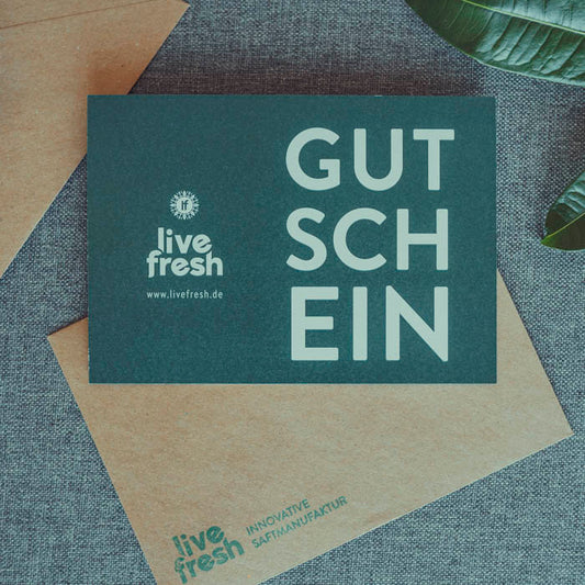 Dunkelgrüne Geschenkkarte mit der Aufschrift „GUTSCHEIN“ in großer, weißer Schrift. Links daneben das Logo und die Website von „LiveFresh“. Die Karte liegt auf einem braunen Umschlag mit weiterem Branding, auf einem grauen Stoffhintergrund.