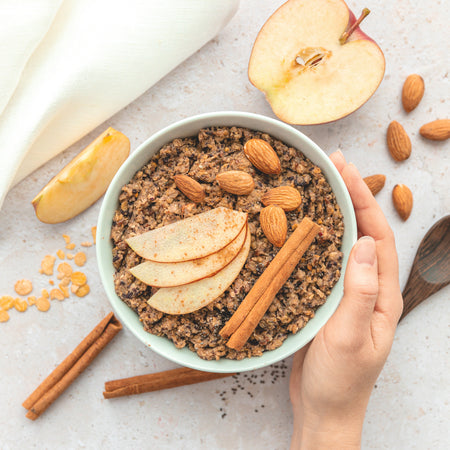 Eine Schüssel Porridge mit Mandeln, Apfelscheiben und Zimtstangen auf hellem Untergrund, umgeben von weiteren Mandeln und Apfelstücken.