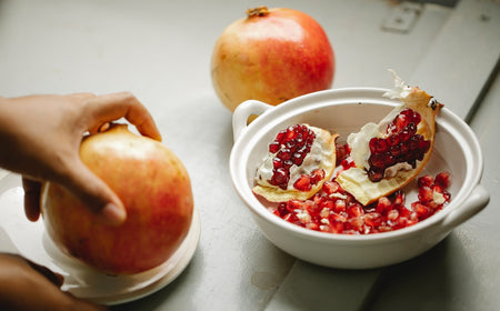 Person schält einen frischen Granatapfel über einer weißen Schüssel, im Hintergrund sind halbierte und ganze Granatäpfel zu sehen.