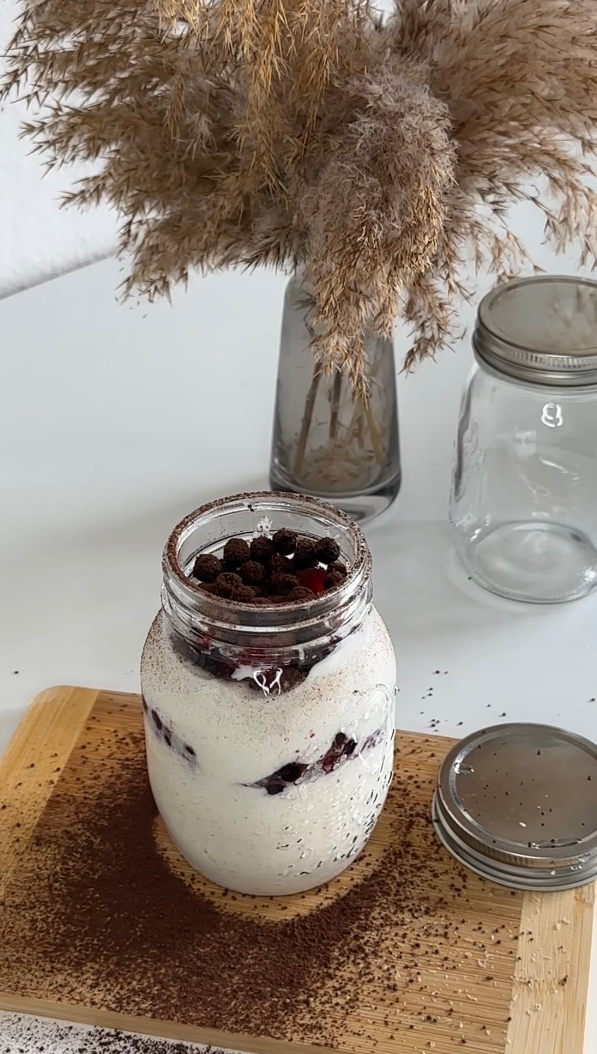 Ein Glas mit cremigem Protein-Grießpudding, geschichtet mit Beeren und getoppt mit Schokocerealien, steht auf einem Holzbrett. Daneben liegen Kakao- und Chiasamen, im Hintergrund stehen Gläser und eine Vase mit Pampasgras.