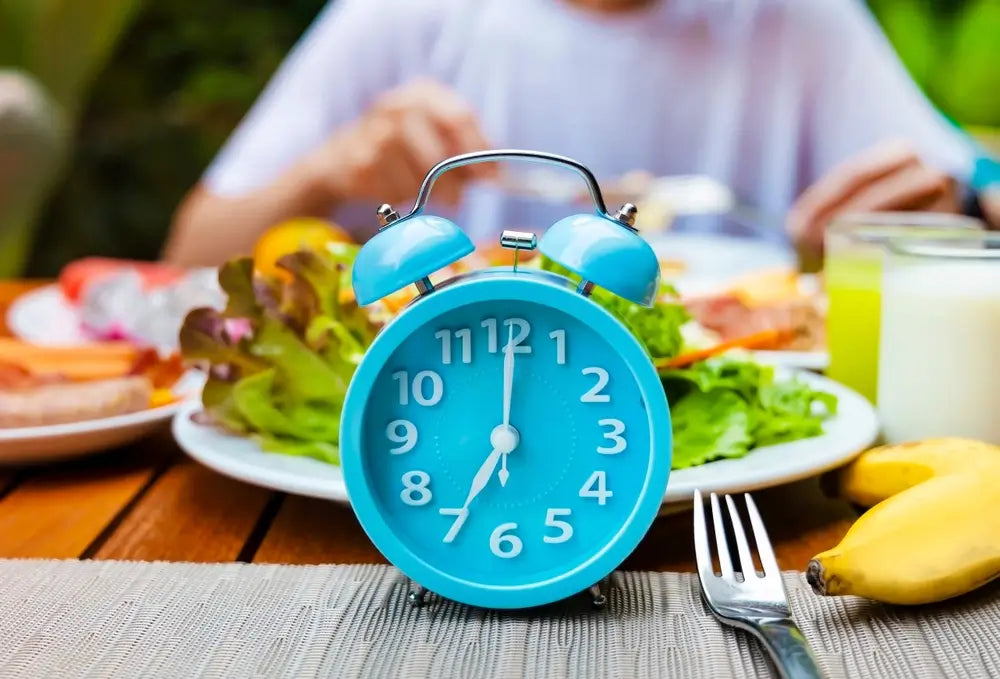 Ein blauer Wecker auf einem Tisch mit frischem Frühstück, darunter Salat, Bananen und Brot, als Symbol für Fasten.