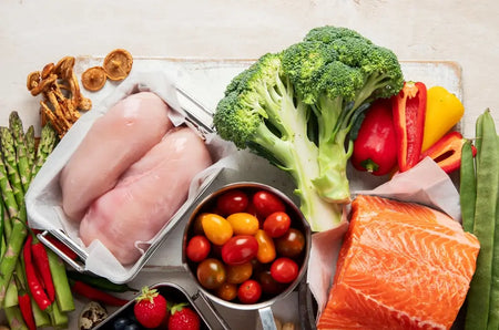 Frische Zutaten für die ketogene Ernährung, darunter Hähnchenbrust, Lachsfilet, Brokkoli, Paprika, Spargel, Pilze und bunte Tomaten, arrangiert auf hellem Untergrund.