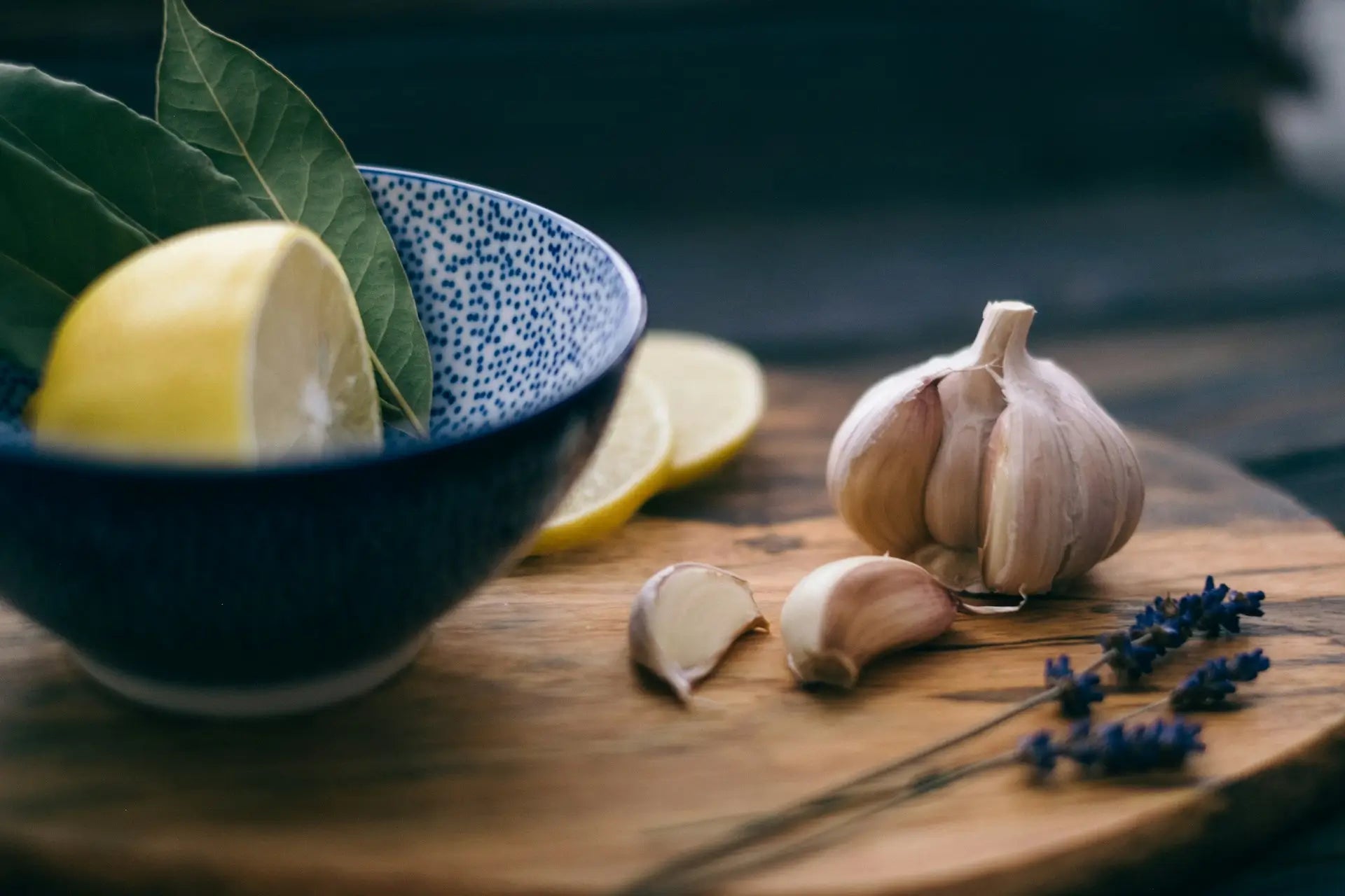 Eine Nahaufnahme einer Holzschneideplatte mit einer Knoblauchknolle, geschnittenen Zitronenscheiben, einem blauen Schälchen und Lavendelzweigen. Die Szene beschreibt eine Zitronen Knoblauch Kur.