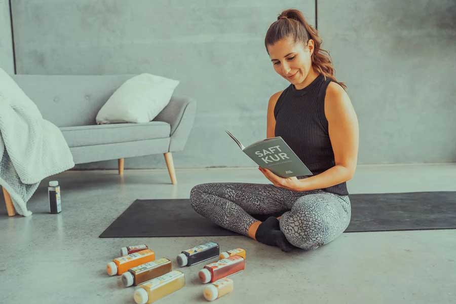 Eine Frau in sportlicher Kleidung, die auf einer Yogamatte sitzt und ein Heft mit der Aufschrift „Saftkur“ liest. Vor ihr auf dem Boden liegen mehrere Flaschen verschiedener Säfte in unterschiedlichen Farben. Im Hintergrund steht ein graues Sofa mit einem Kissen und einer Decke.