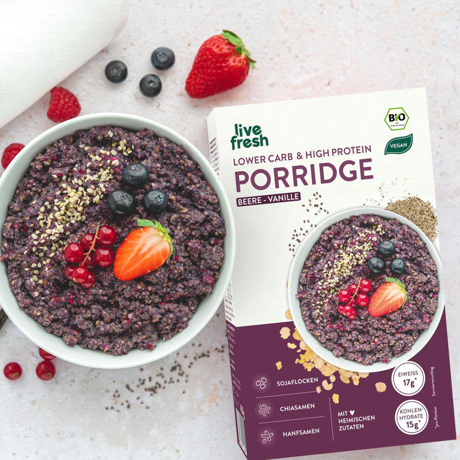 Schüssel „Live Fresh Porridge“ in der Geschmacksrichtung Beere-Vanille, garniert mit frischen Beeren wie Erdbeeren, Johannisbeeren und Blaubeeren sowie Hanfsamen. Daneben steht die Verpackung des Porridges, die ebenfalls Beeren und Vanille als Geschmacksprofil hervorhebt. Die Verpackung betont, dass das Porridge „Lower Carb & High Protein“ ist, vegan und aus heimischen Zutaten wie Sojaflocken, Chiasamen und Hanfsamen besteht, mit 17g Eiweiß und 15g Kohlenhydraten pro Portion.