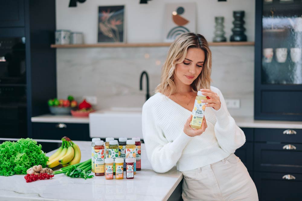 Eine Frau in einer modernen Küche, die eine Flasche „Live Fresh“-Saft in der Hand hält und das Etikett betrachtet. Vor ihr auf der Arbeitsfläche stehen weitere Flaschen mit verschiedenen Saftsorten. Auf der Arbeitsplatte liegen auch frische Zutaten wie Salat, Bananen, Ingwer und Frühlingszwiebeln, die eine gesunde Ernährung symbolisieren