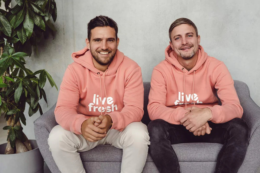 Zwei Männer die in korallenfarbenen Hoodies mit dem Logo "Live Fresh", entspannt auf einem grauen Sofa sitzen. Sie lächeln freundlich in die Kamera, was eine sympathische und zugängliche Atmosphäre vermittelt. Die große Pflanze im Hintergrund verstärkt den natürlichen, frischen Eindruck, passend zur Marke "Live Fresh".