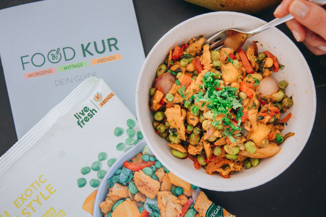 Eine bunte Gemüse-Bowl mit Erbsen, Paprika, Zwiebeln und Kräutern. Daneben liegt die Verpackung des „Exotic Style“ Gerichts von Live Fresh und ein „Food Kur“ Guide. Es vermittelt gesunde Ernährung und wirkt inspirierend für einen strukturierten, nahrhaften Ernährungsplan.