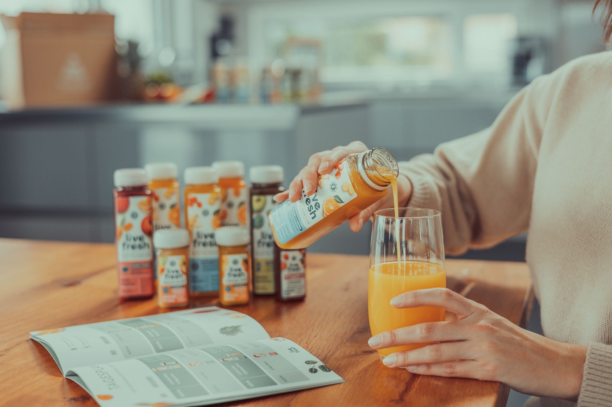 Eine Person, die einen Live Fresh Saft in ein Glas gießt, während mehrere weitere Flaschen des Saftes und ein Guide auf dem Tisch zu sehen sind. Die Szene wirkt ruhig und gesundheitsbewusst, passend zu einer strukturierten Saftkur oder Ernährungsumstellung.