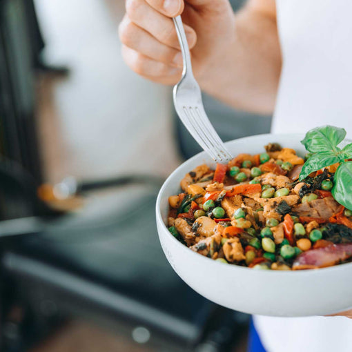 Nahaufnahme einer bunten Gemüse-Bowl mit Erbsen, Paprika, Hülsenfrüchten und frischem Basilikum, die vor einem Fitnessgerät in der Hand gehalten wird.