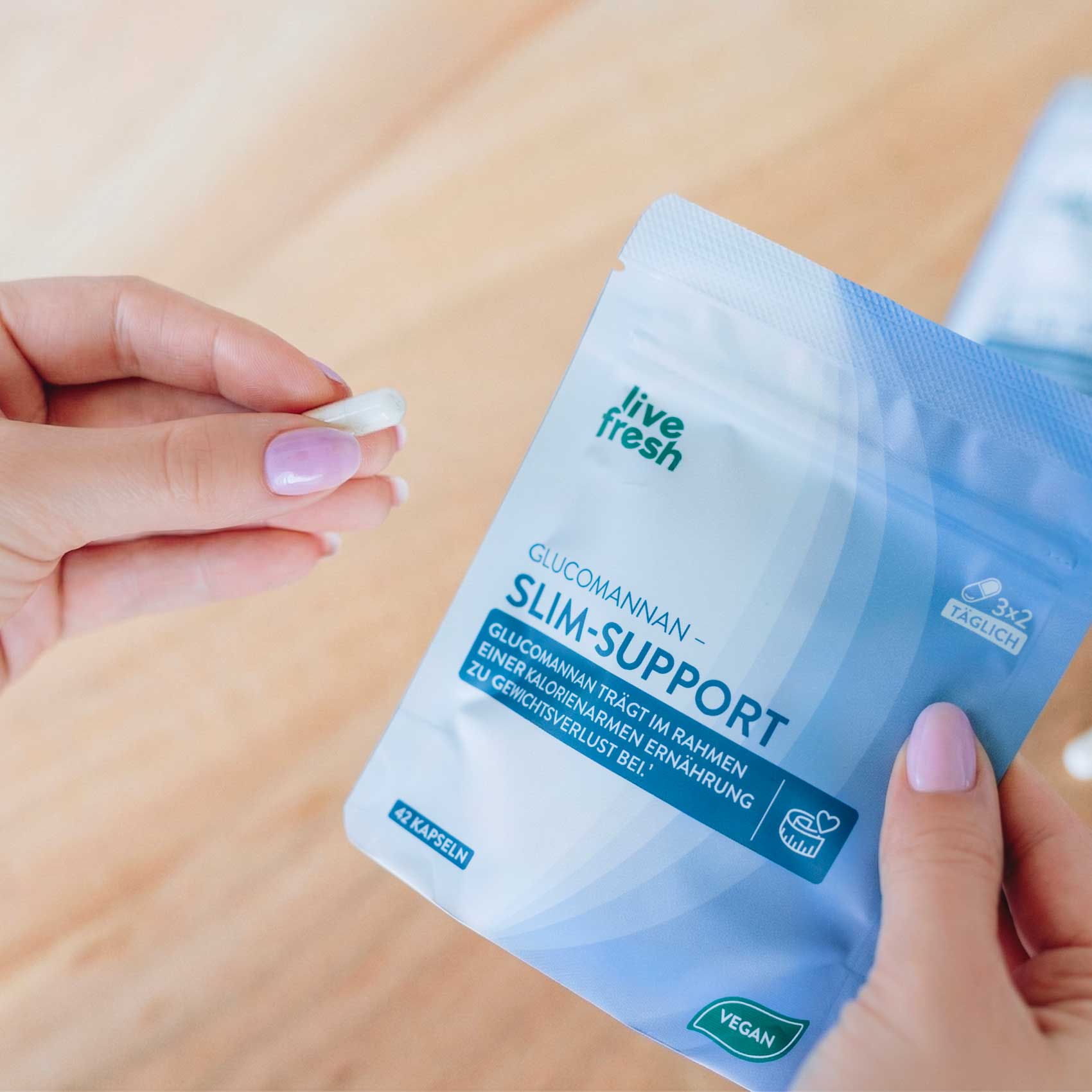 Eine Hand hält eine einzelne Kapsel neben einem Beutel „live fresh Glucomannan Slim-Support“, der teilweise geöffnet ist. Auf der Verpackung steht „GLUCOMANNAN – SLIM-SUPPORT“, „42 KAPSELN“ und „VEGAN“, während die Szene die Einnahme des Produkts im Alltag zeigt.