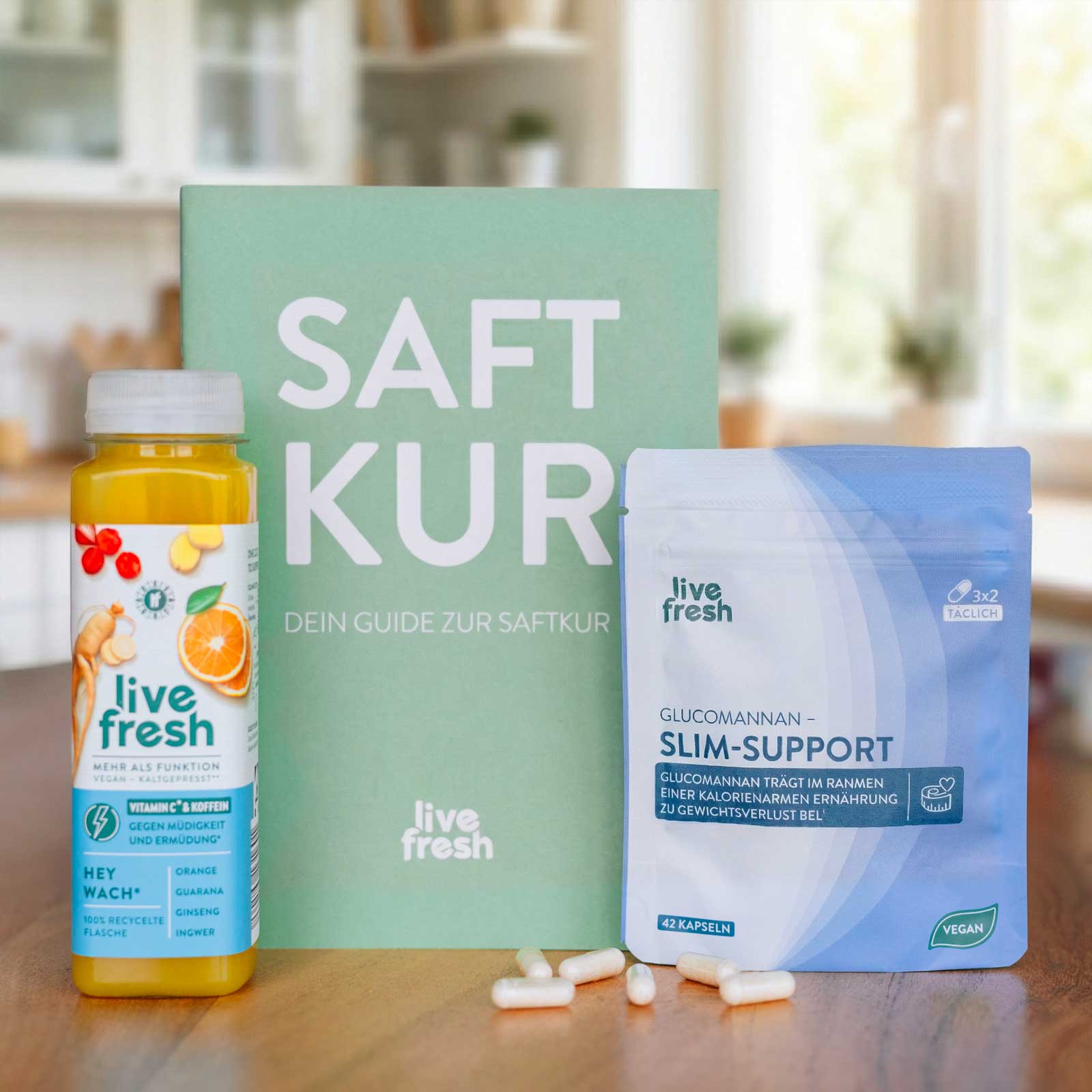 Productset met een fles “levend vers” sap, een boek met de titel “SAFT KUR” en een zakje “Glucomannan Slim-Support” staat op een tafel, met verschillende capsules ervoor. De tekst “GLUCOMANNAN – SLIM SUPPORT” en “GLUCOMANNAN DRAAGT BIJ AAN GEWICHTSVERLIES ALS ONDERDEEL VAN EEN LAAG CALORIE DIEET” zijn duidelijk zichtbaar op de verpakking.