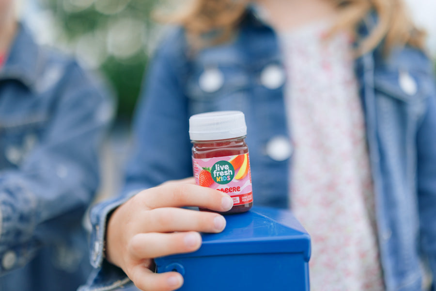Close-up van een LiveFresh Kids Shot met aardbeiensmaak, met op de achtergrond een meisje in een spijkerjasje.