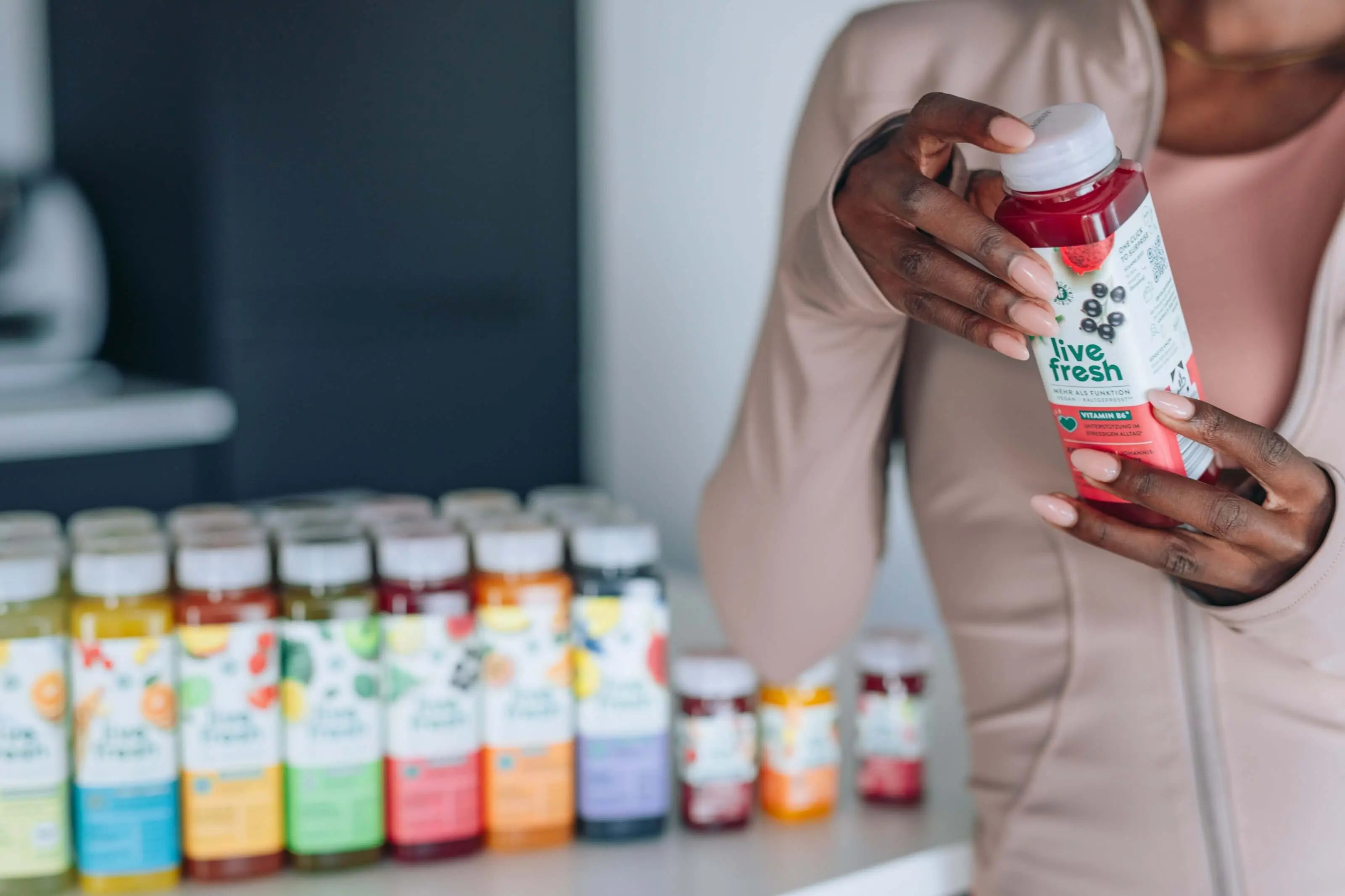 Person hält eine Flasche „Live Fresh“-Saft in der Hand, im Hintergrund stehen weitere bunte Saftflaschen aufgereiht auf einer Theke.