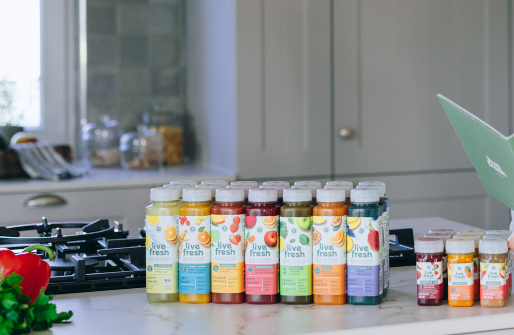 Mehrere bunte LiveFresh-Saftflaschen und Shots in verschiedenen Sorten (Happy Lemon, Hey Wach, Sommer Gefühle, Rote Rakete, Grüne Gefühle, Gold Rausch, Keep Cool) stehen aufgereiht auf einer hellen Küchenarbeitsplatte neben einem Herd. Im Hintergrund ein modernes Küchen-Setting.