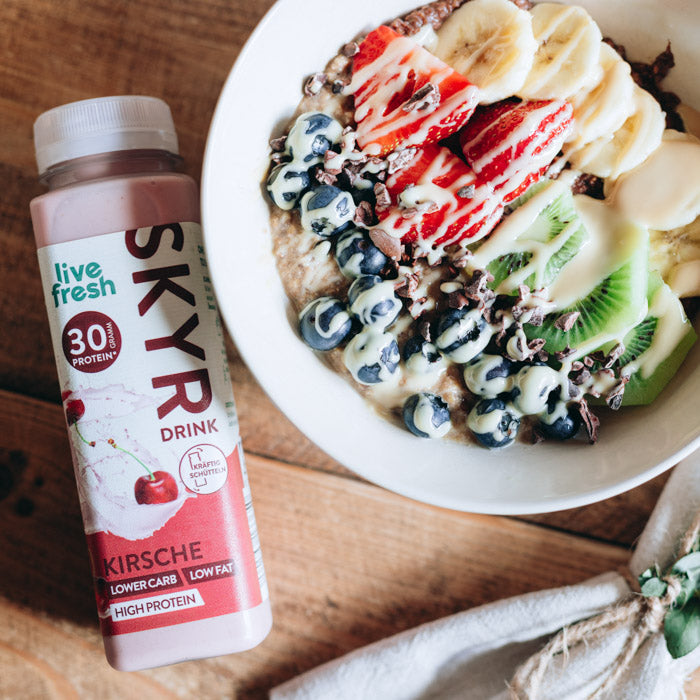 Eine Schüssel mit Joghurt, garniert mit Bananenscheiben, Erdbeeren, Kiwi, Blaubeeren und Kakaonibs. Daneben steht eine Flasche „Live Fresh Skyr Drink Kirsche“ mit 30g Protein. Die Flasche wirbt mit einem niedrigen Kohlenhydratgehalt, fettarm und proteinreich, und unterstreicht eine gesunde und nahrhafte Mahlzeit.