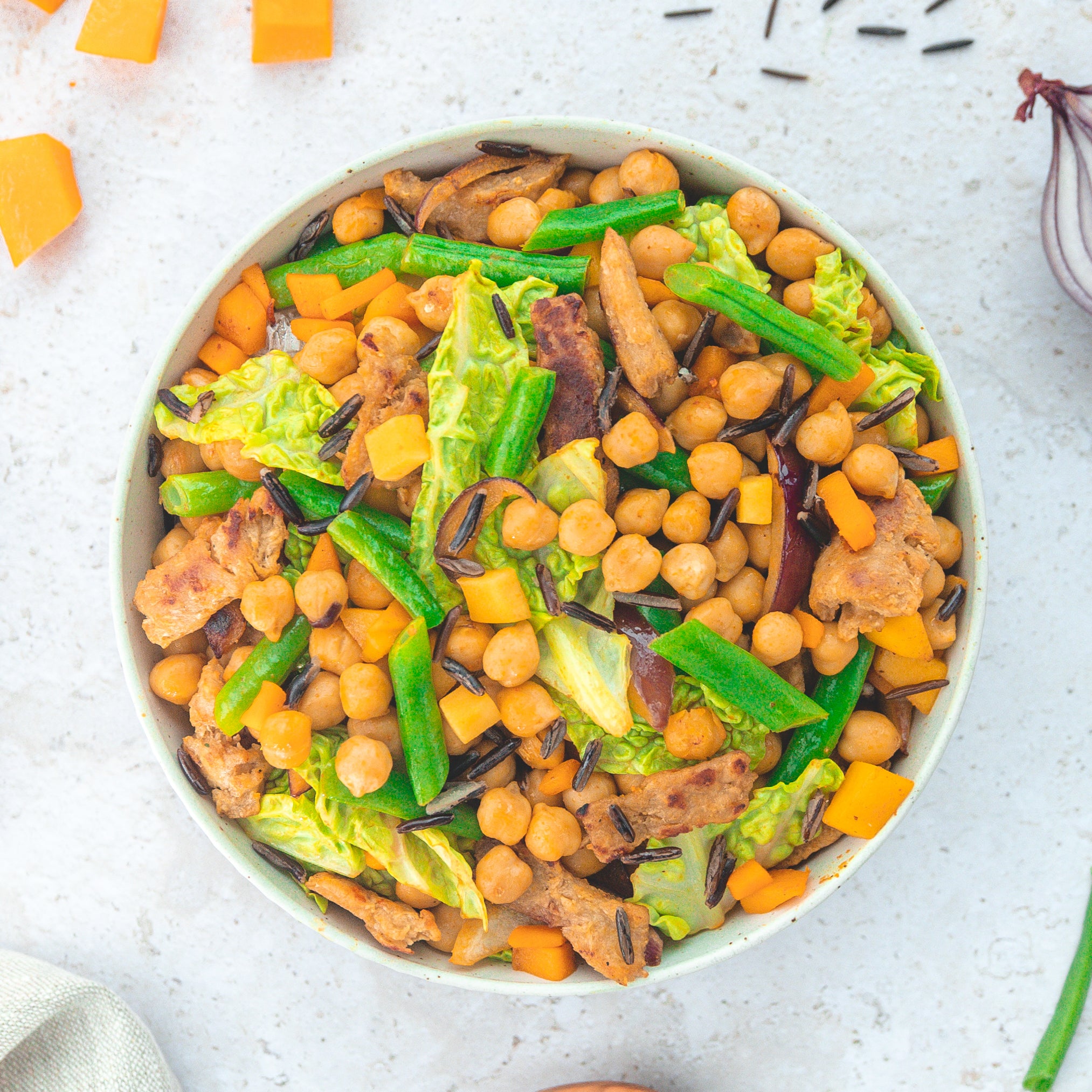 Eine bunte Bowl, gefüllt mit Kichererbsen, grünen Bohnen, Wildreis, Kürbiswürfeln, Salat und Fleischersatzstreifen. Die farbenfrohe Mischung suggeriert eine gesunde, vegane Mahlzeit mit viel Gemüse und pflanzlichen Proteinen.