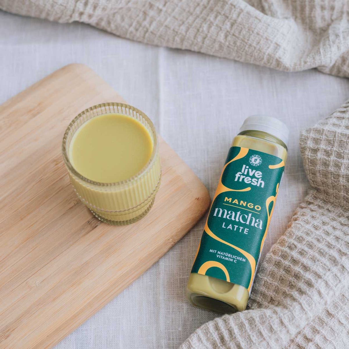„Live Fresh“ Mango Matcha Latte in Flasche neben gefülltem Glas auf Holzbrett, arrangiert auf hellem Stoff mit natürlichem Licht.
