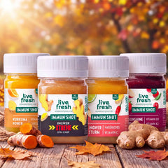 Vier Live Fresh Immunshots in den Sorten Kurkuma Power, Ingwer Xtreme, Ingwer Sturm und Sunshine Berry stehen auf einem Holztisch mit frischem Ingwer, Kurkuma und bunten Herbstblättern. Die verschiedenen Shots präsentieren die Sortenauswahl für ein Immunshots Monatspaket.
