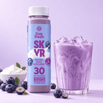 Live Fresh Skyr Protein Drink Heidelbeere neben einem Glas mit eisgekühltem cremigem Heidelbeerdrink. Auf der Verpackung steht "live fresh SKYR PROTEIN DRINK HEIDELBEERE MIT KOLLAGEN 30 GR. PROTEIN HIGH PROTEIN LOWER CARB NO ADDED SUGAR". Frische Heidelbeeren und eine Schale mit cremigem Skyr stehen daneben.