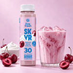 Live Fresh Skyr Protein Drink Kirsche neben einem Glas mit eisgekühltem rosafarbenem Kirschdrink. Auf der Verpackung steht "live fresh SKYR PROTEIN DRINK KIRSCHE MIT KOLLAGEN 30 GR. PROTEIN HIGH PROTEIN LOWER CARB NO ADDED SUGAR". Frische und halbierte Kirschen sowie eine Schale mit cremigem Skyr stehen daneben.