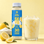 Live Fresh Skyr Protein Drink Zitrone neben einem Glas mit eisgekühltem cremigem Zitronendrink. Auf der Verpackung steht "live fresh SKYR PROTEIN DRINK ZITRONE MIT KOLLAGEN 30 g PROTEIN HIGH PROTEIN NO ADDED SUGAR". Zitronen und eine Schale mit cremigem Skyr stehen daneben.