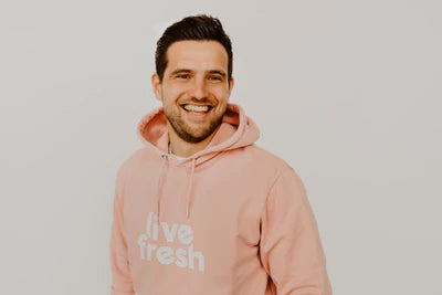 Das Bild zeigt einen lächelnden Mann in einem rosa "Live Fresh" Hoodie vor einem hellen Hintergrund. Er wirkt sympathisch und entspannt, was eine freundliche und nahbare Atmosphäre vermittelt.