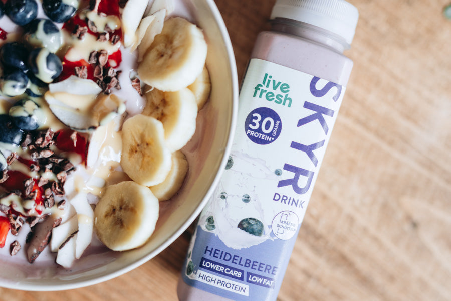 Flasche „Live Fresh Skyr Drink“ in der Geschmacksrichtung Heidelbeere, die neben einer Schüssel mit frischem Obst liegt. In der Schüssel sind Bananenscheiben, Heidelbeeren, Erdbeeren und weitere Zutaten wie Kakaonibs und Joghurt zu sehen. Der Skyr Drink betont 30g Protein, „Low Carb“ und „Low Fat“.