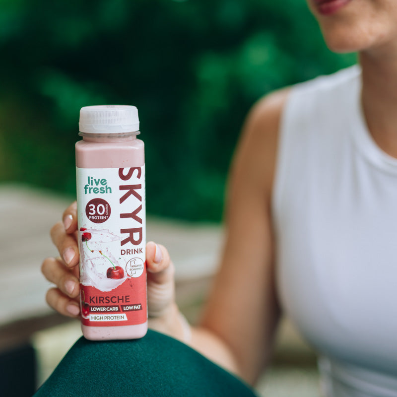 Eine Person, die eine Flasche „Live Fresh Skyr Drink Kirsche“ in der Hand hält. Die Flasche hebt 30g Protein, niedrigen Kohlenhydratgehalt, fettarm und proteinreich hervor. Die Person trägt ein weißes Oberteil und sitzt draußen, was auf einen aktiven und gesunden Lebensstil hinweist.