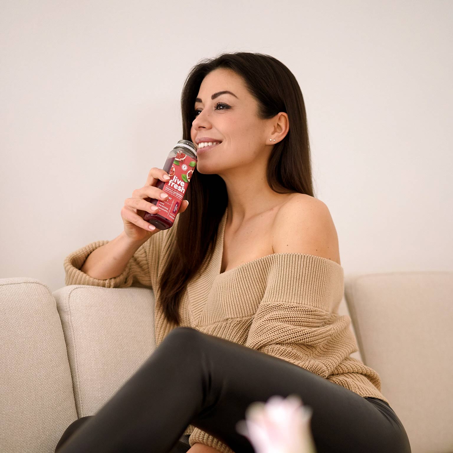 Eine Frau, die auf einem hellen Sofa sitzt und aus einer Saftflasche trinkt. Auf der Flasche steht "Live Fresh" mit Abbildungen von Früchten, was darauf hindeutet, dass es sich um einen frischen Saft oder Smoothie handelt. Sie trägt einen beigefarbenen, schulterfreien Pullover und schwarze Hosen, während sie lächelnd ihr Getränk genießt.