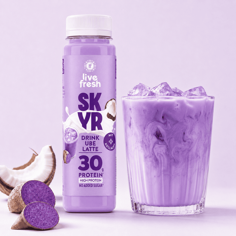 Live Fresh Skyr Drink Ube Latte neben einem Glas mit eisgekühltem lilafarbenem Ube Drink. Auf der Verpackung steht "live fresh SKYR DRINK UBE LATTE MIT KOLLAGEN 30 g PROTEIN HIGH PROTEIN NO ADDED SUGAR". Aufgeschnittene Ube und Kokosnuss liegen daneben.