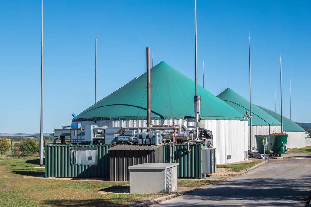 Biogasanlage mit mehreren großen Tanks, die grüne, kuppelförmige Dächer haben. Im Vordergrund sind technische Geräte und Rohre zu sehen, die zur Energieerzeugung beitragen. Der klare Himmel und die weitläufige Landschaft im Hintergrund unterstreichen die umweltfreundliche Energieproduktion.