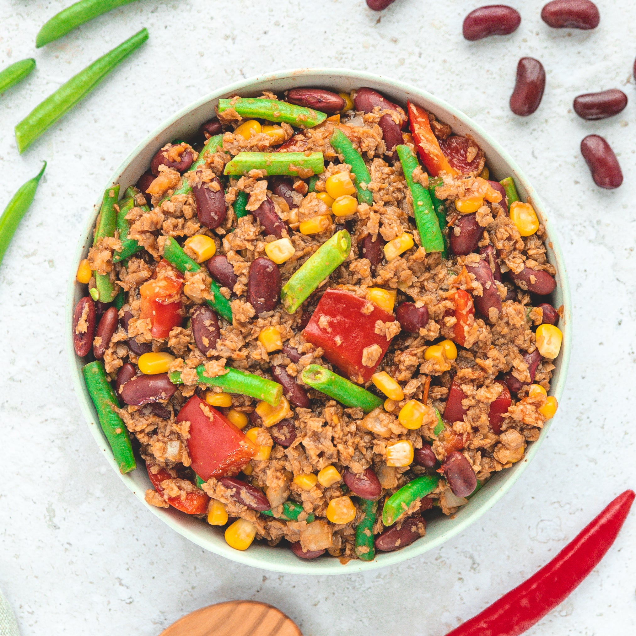 Eine Schale mit "Chili sin Carne" von Live Fresh. Die vegane Mahlzeit enthält grüne Bohnen, Kidneybohnen, Mais, Tomaten und pflanzliches Hack. Um die Schale herum liegen frische Chilischoten und Bohnen, was die Zutaten des Gerichts hervorhebt.