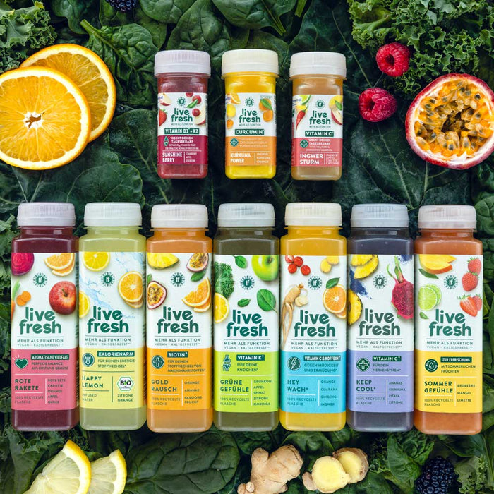 Produkte von LiveFresh: Oben drei Shots – Sunshine Berry, Kurkuma Power und Ingwer Sturm. Darunter sieben verschiedene kaltgepresste Säfte – Rote Rakete, Happy Lemon, Gold Rausch, Grüne Gefühle, Hey Wach, Keep Cool und Sommergefühle. Im Hintergrund Blattgrün, Orangen, Passionsfrucht und Himbeeren.