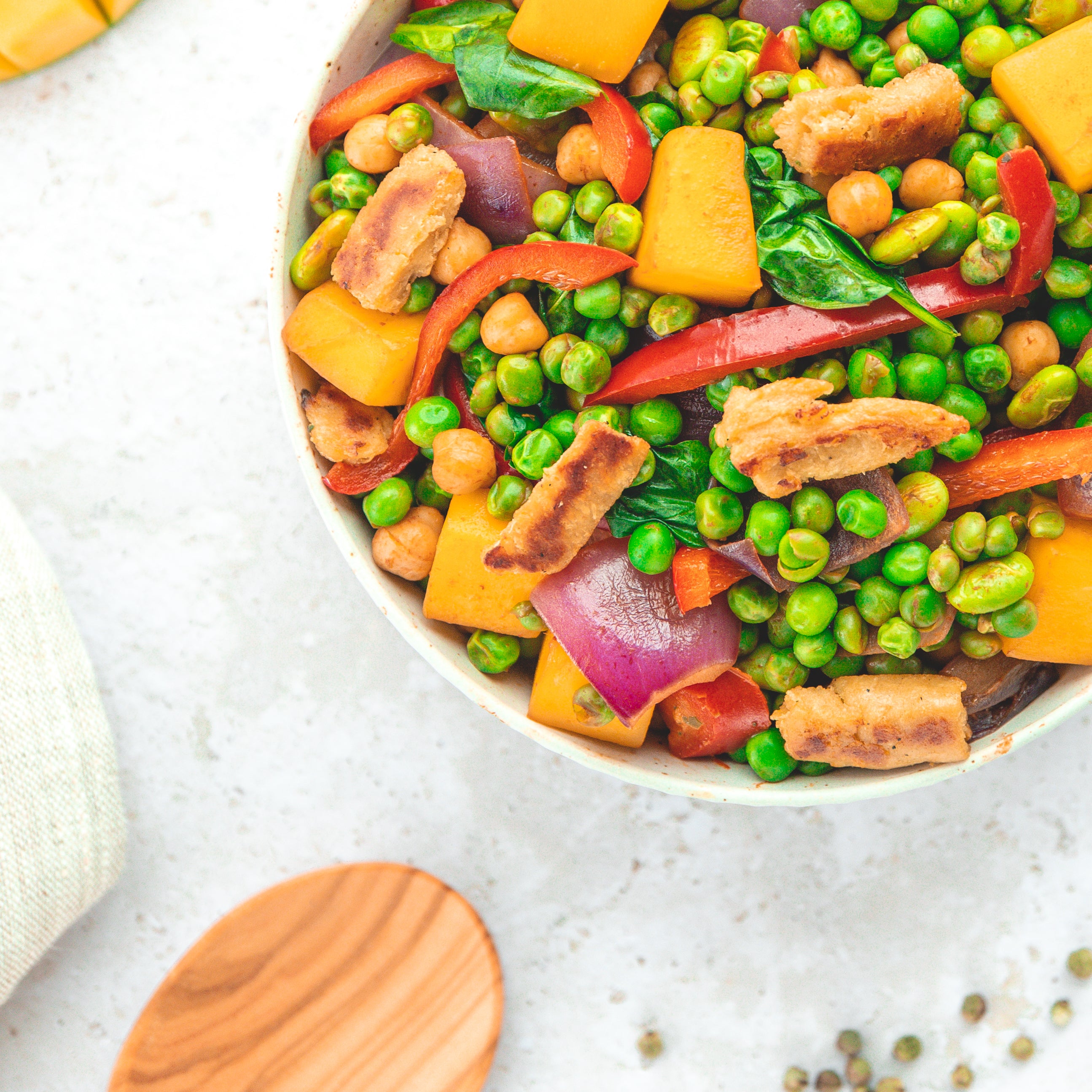 Eine exotische Bowl mit buntem Gemüse. Enthalten sind grüne Erbsen, Paprikastreifen, rote Zwiebeln, Kichererbsen, Spinat und leuchtend gelbe Mangostücke. Ergänzt wird das Gericht durch vegane Proteinstücke. Die Farben und Vielfalt der Zutaten lassen es besonders frisch und nahrhaft wirken.