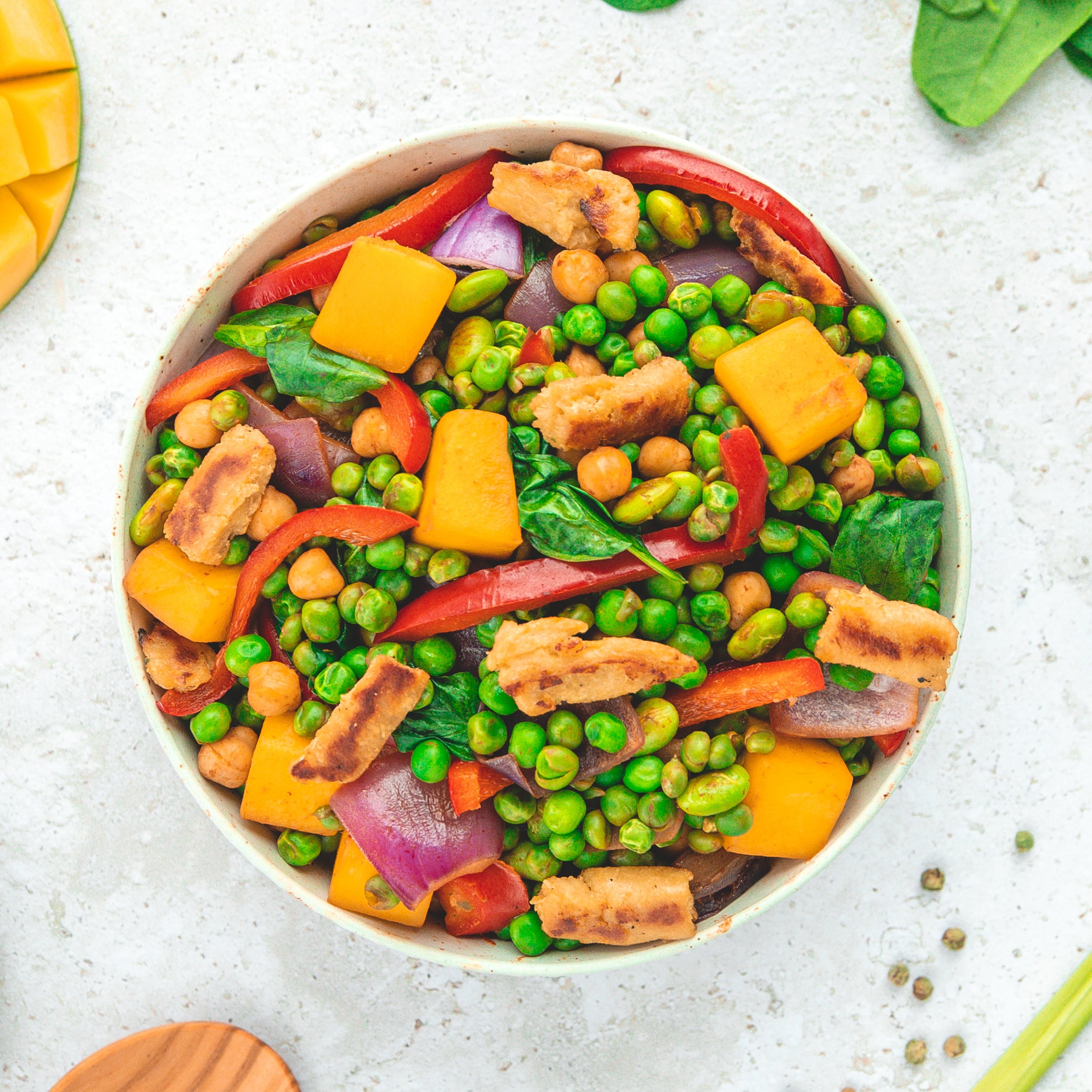 Eine bunte Bowl mit frischen Zutaten wie grünen Erbsen, roten Paprikastreifen, roten Zwiebeln, Kichererbsen, Spinat und goldgelben Mangostücken, ergänzt durch knusprige vegane Proteinstücke. Die Mischung vermittelt ein gesundes, ausgewogenes und farbenfrohes Gericht.