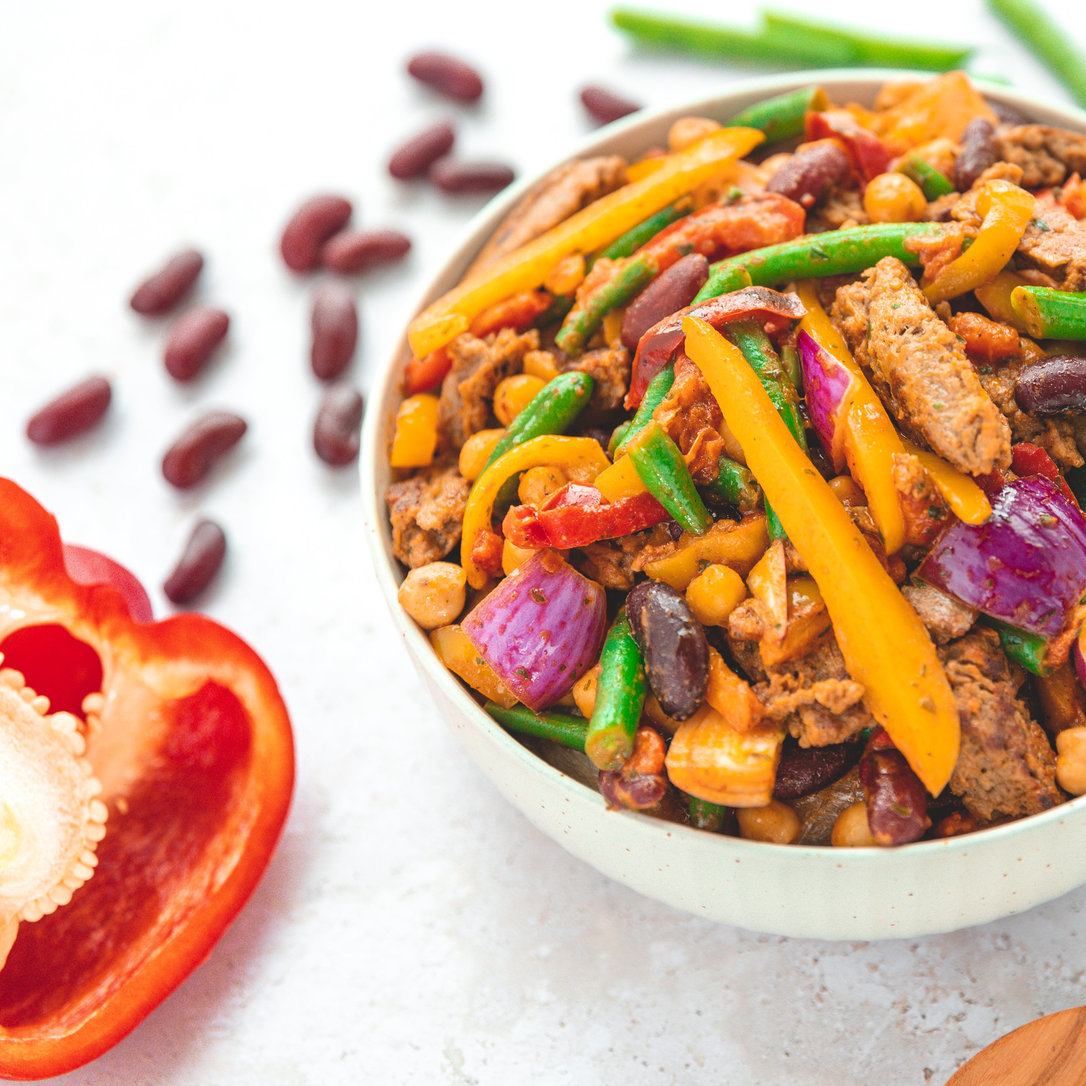 Nahaufnahme einer Fajita Bowl, gefüllt mit buntem, gebratenem Gemüse wie gelber Paprika, roten Zwiebeln, grünen Bohnen und Kidneybohnen, kombiniert mit veganem Fleischersatz und Kichererbsen. Im Hintergrund sind frische rote Kidneybohnen und eine aufgeschnittene Paprika zu sehen, die die Frische und Vielfalt der Zutaten betonen.