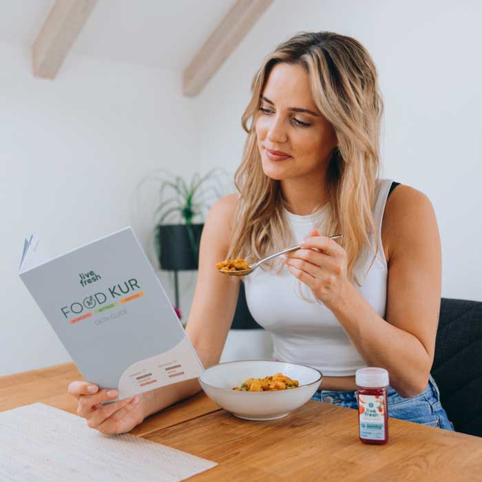 Eine Frau, die an einem Tisch sitzt und eine Schüssel Essen in der Hand hält, während sie in einem Heft mit der Aufschrift „Food Kur“ blättert. Sie trägt ein weißes Top und lächelt leicht. Neben ihr auf dem Tisch steht eine kleine Flasche „Live Fresh“-Saft. Die Szene vermittelt den Eindruck einer ausgewogenen Ernährung im Rahmen einer Food-Kur